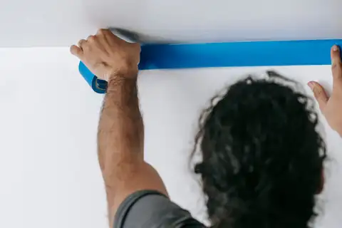 Person seen from behind pressing blue painter's tape along a ceiling line